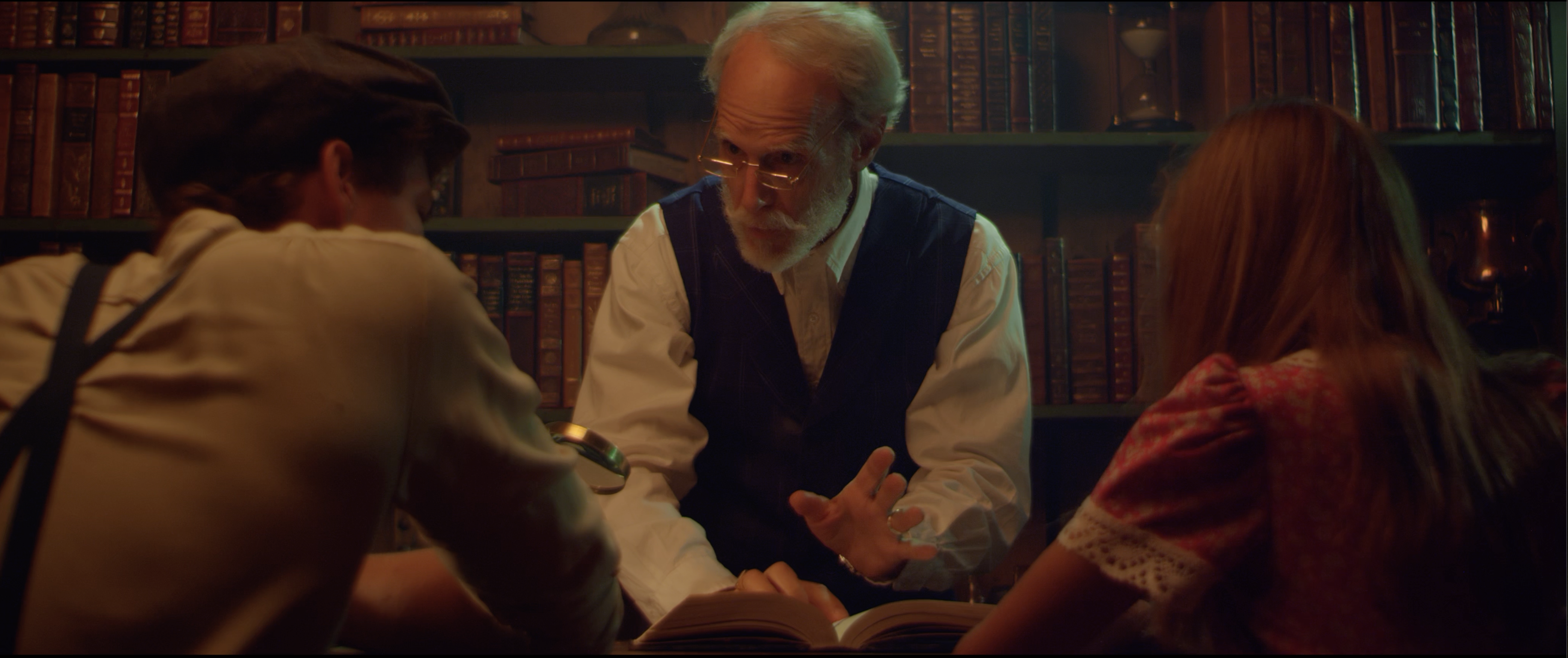 An older man stands in front of a bookshelf talking to two people whose backs are to the camera