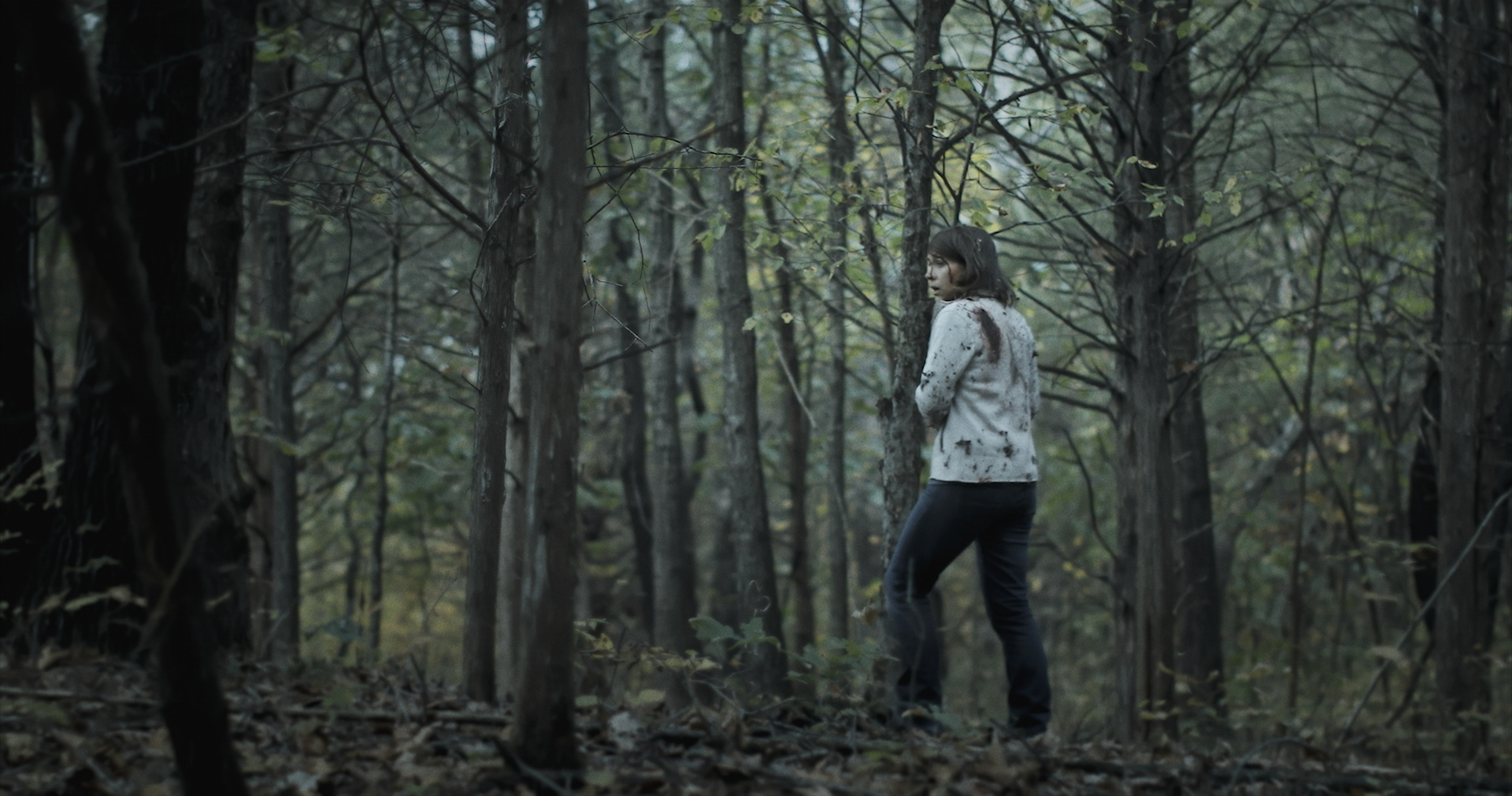 A woman with a white sweater covered in dirt walks through a pine forest